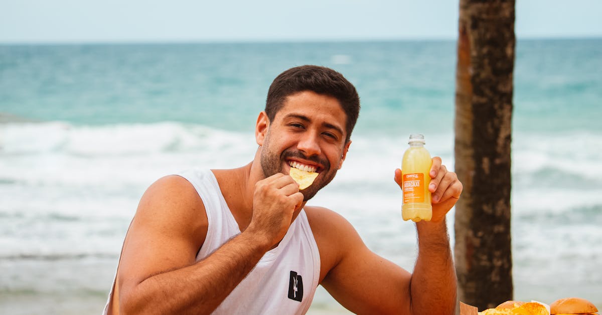 Man in a white tank top enjoys chips and juice by the seaside, smiling.