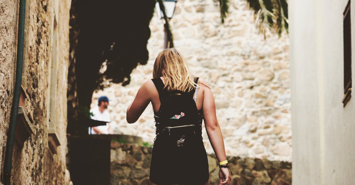 Blonde woman strolls through a picturesque stone alleyway on a sunny day, wearing a fashionable black outfit.