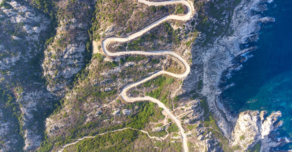 Aerial image of a winding road traversing rocky coastal terrain by a clear blue sea.