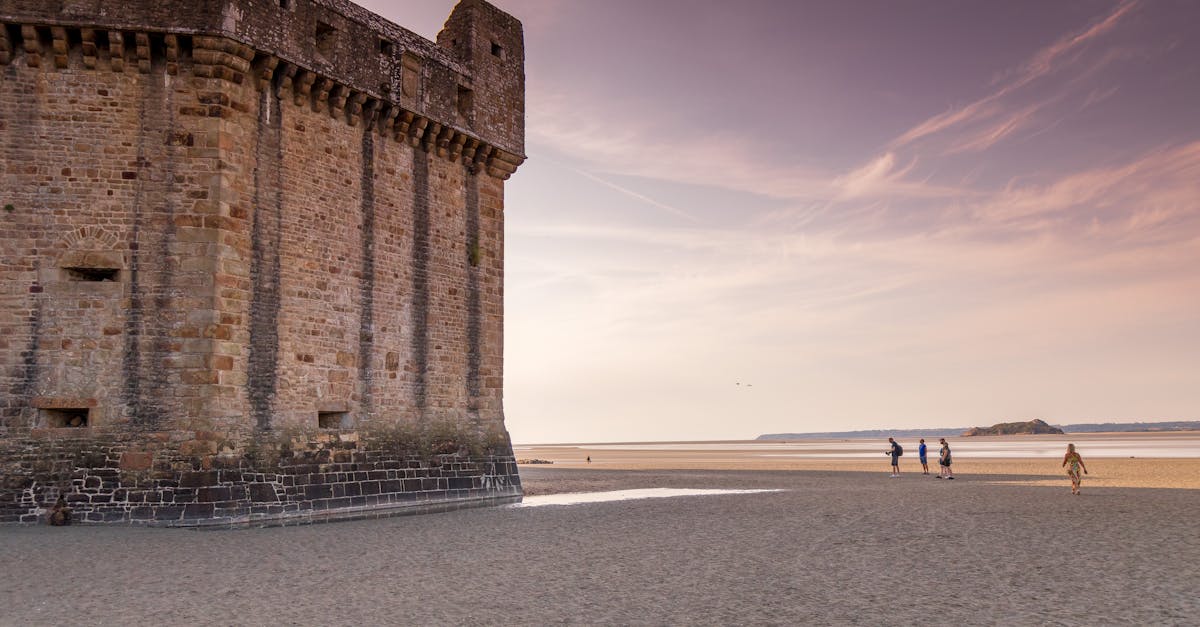 Scenic view of Le Mont-Saint-Michel's ancient fortress with people walking on the beach at sunset.