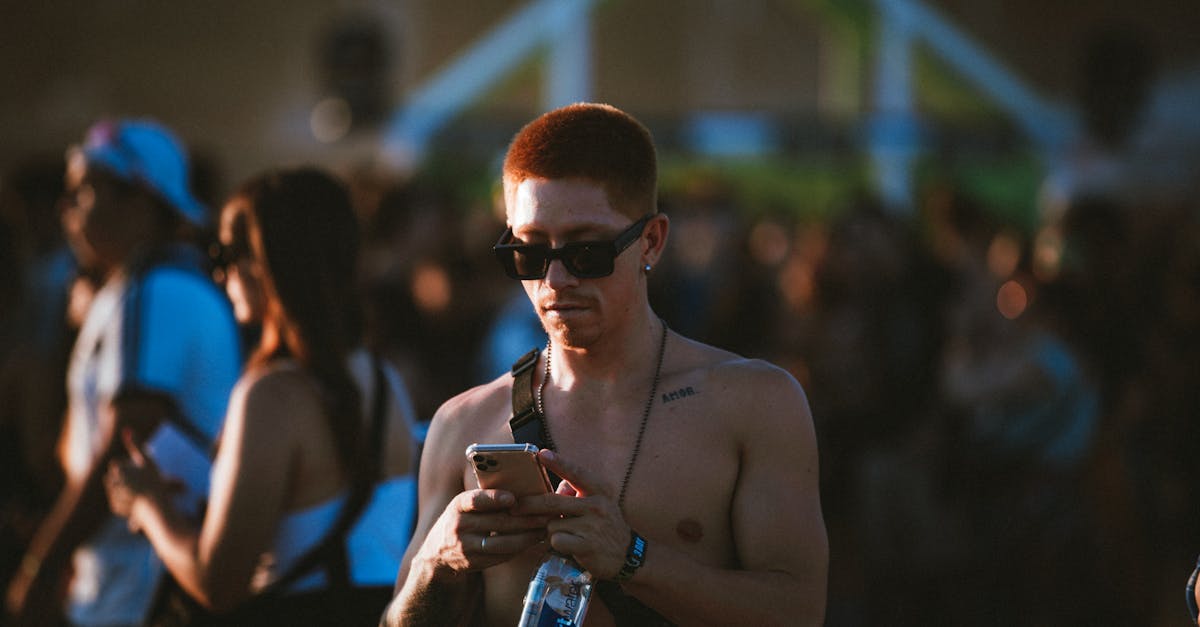 Shirtless man wearing sunglasses at a lively outdoor music festival, checking phone.