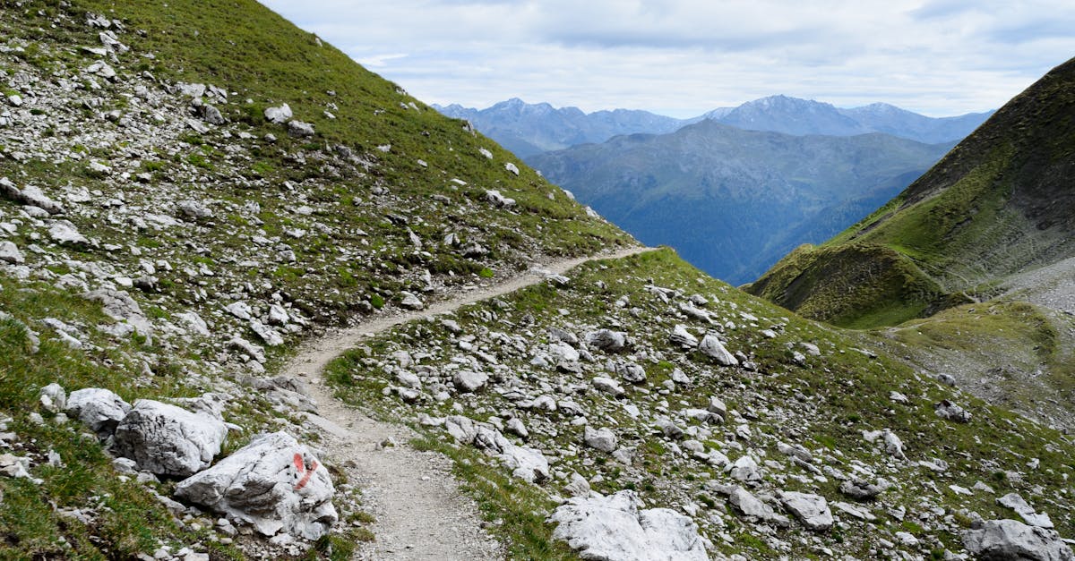 A winding trail through a rocky, lush green mountain landscape under a cloudy sky.