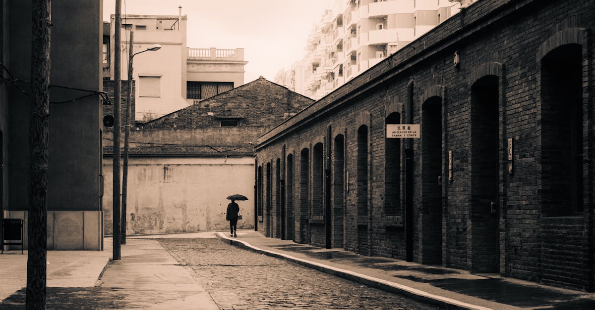 Explore the charm of a quiet cobblestone street in Barcelona with a solitary figure under an umbrella. Timeless and atmospheric.