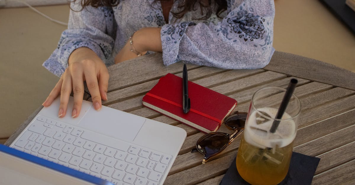 A woman works on a laptop at an outdoor café table with a drink, notebook, and sunglasses.