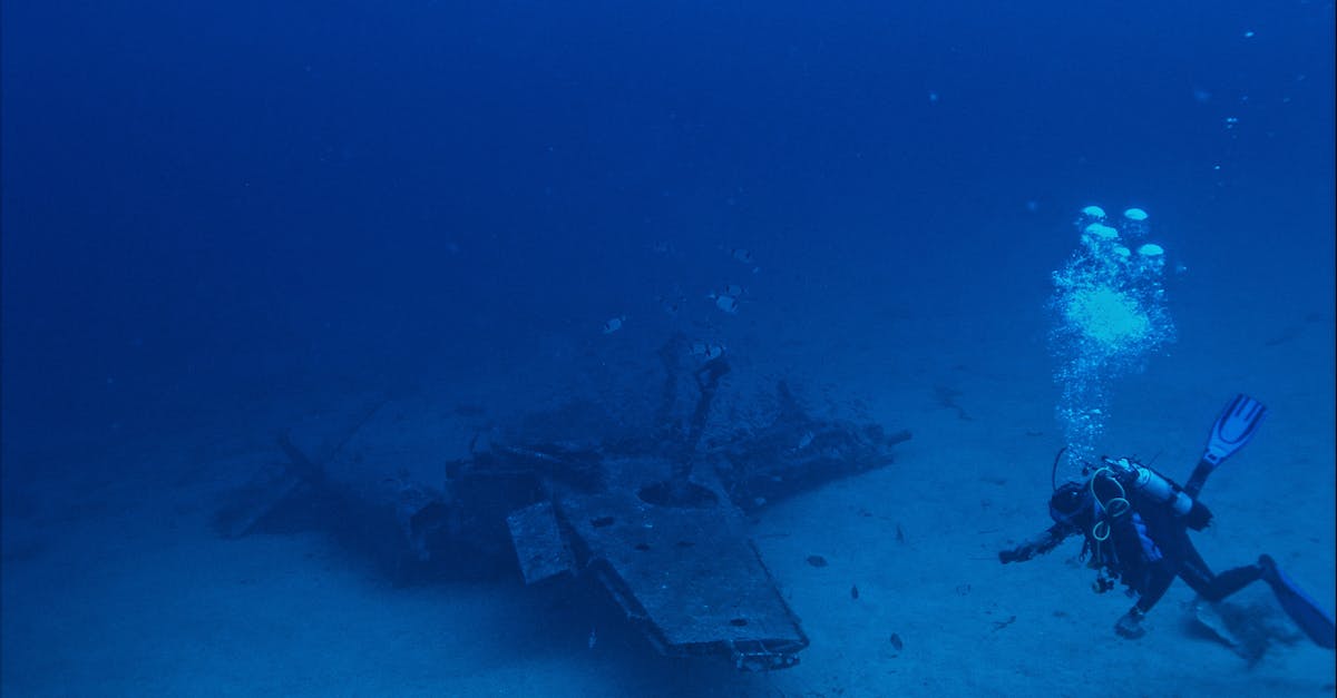 Unrecognizable person with oxygen tank emitting bubbles and swimming towards remains of sunken plane in blue sea water