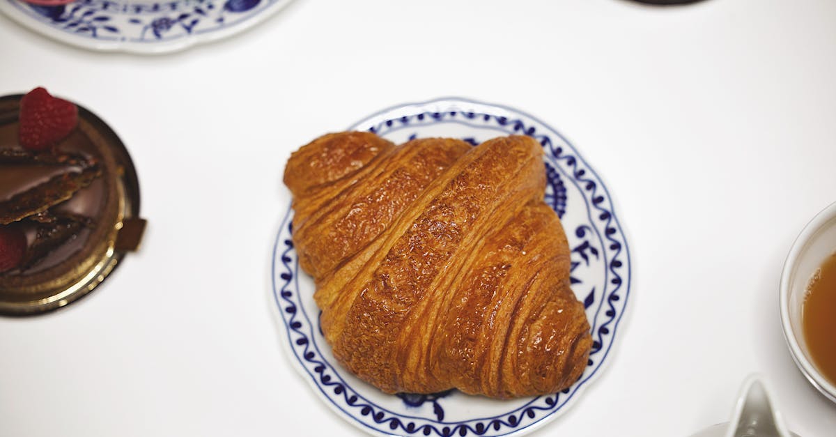A perfectly baked croissant on a blue patterned plate surrounded by desserts and tea.