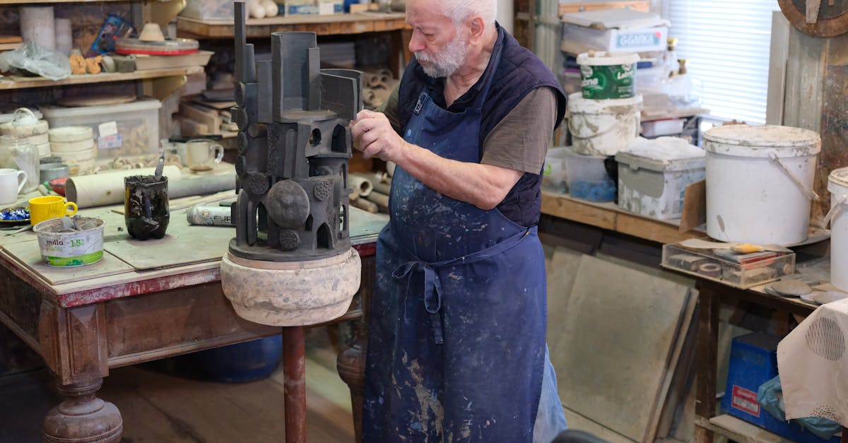 A senior male artist sculpting a clay ceramic piece in his cluttered workshop filled with pottery tools.