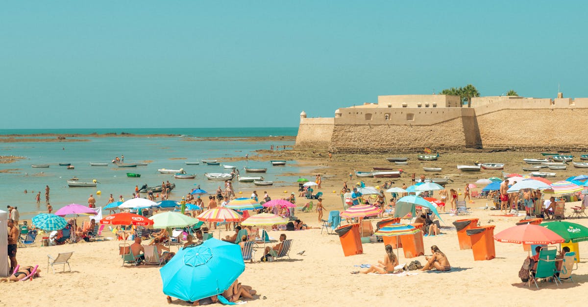 A vibrant beach scene with colorful umbrellas near an historic coastal fort under a bright summer sky.
