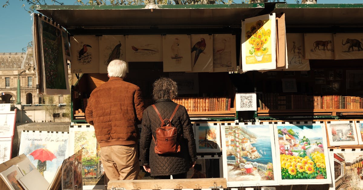 A couple explores an art stall in Paris under the sun, displaying vibrant paintings.