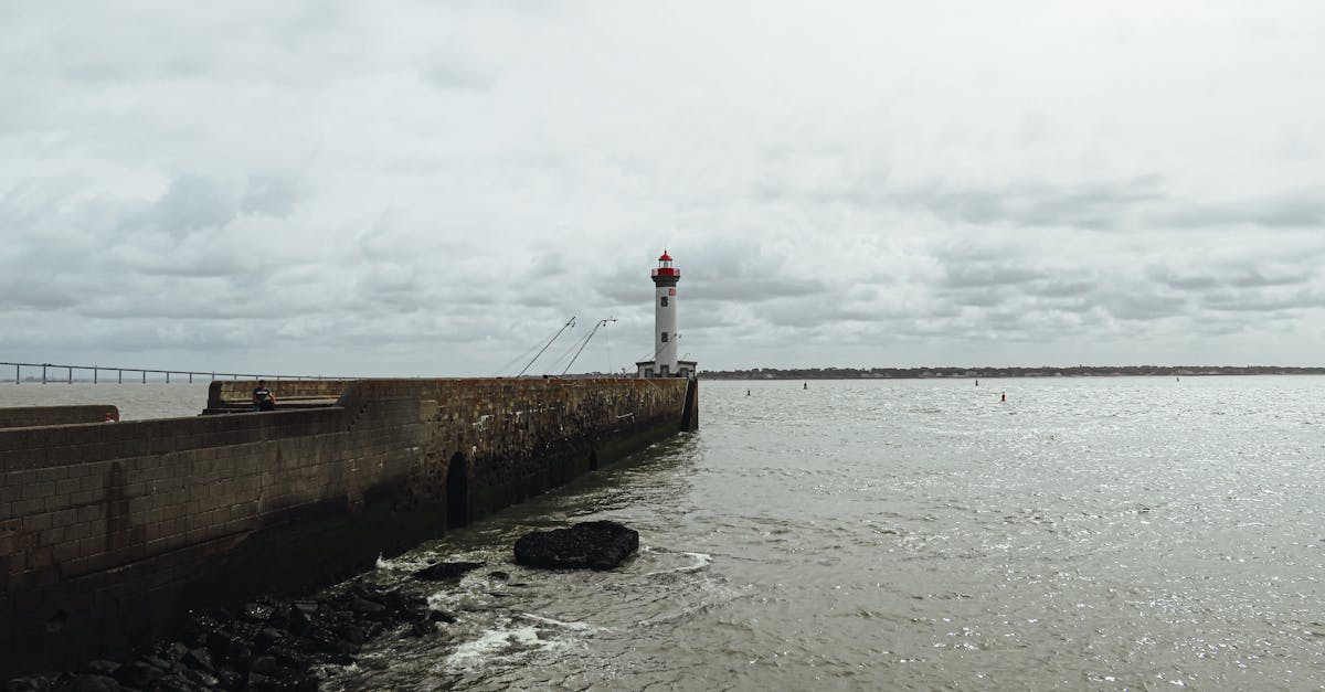 A coastal lighthouse stands at the end of a long pier on an overcast day.