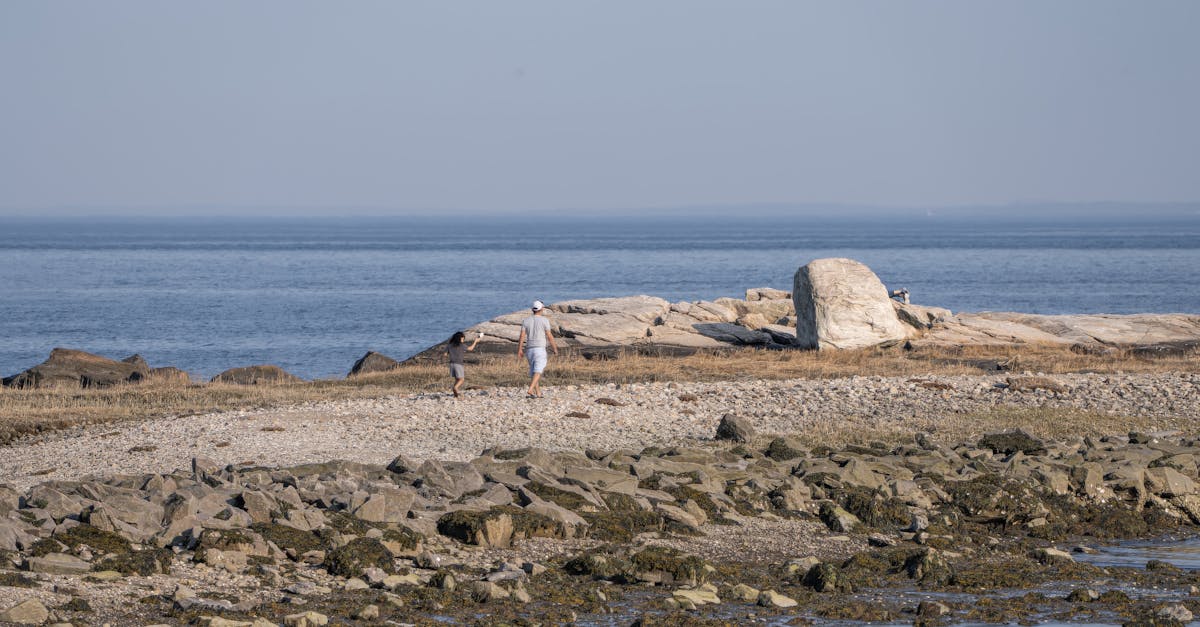 Two people enjoy a walk on a rocky beach with distant ocean view, clear sky, and serene atmosphere.