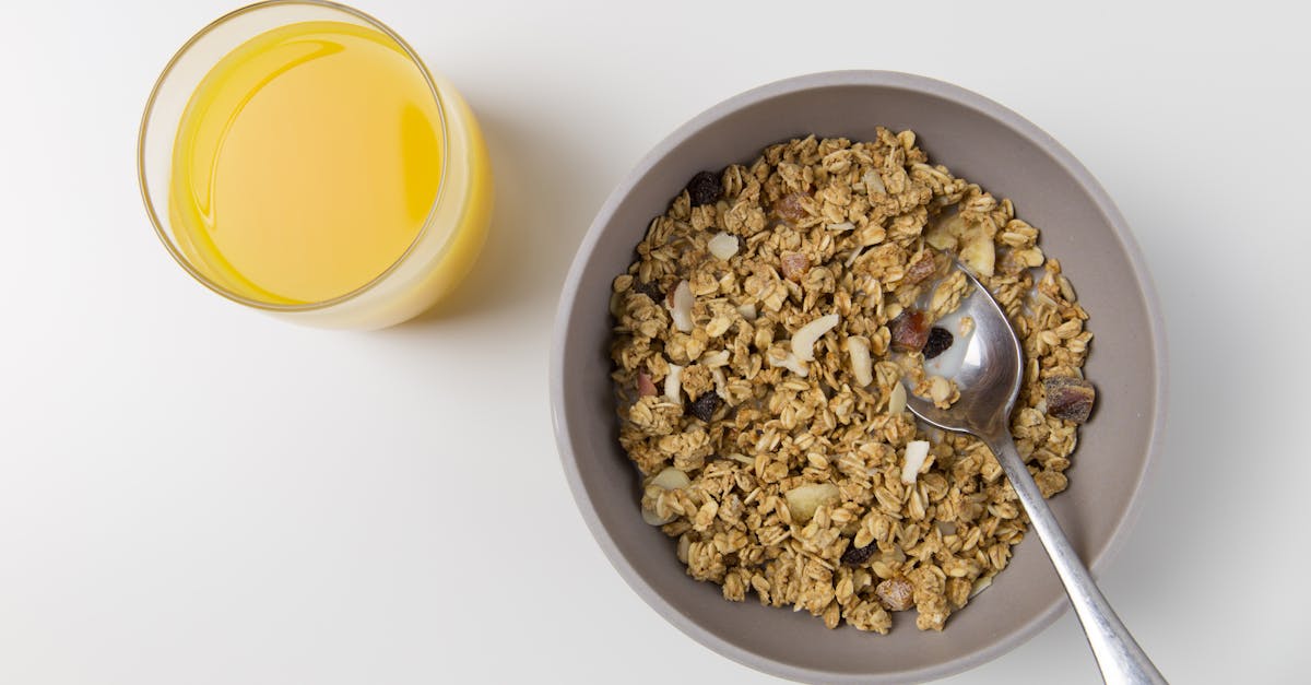 A top view of granola in a bowl with a spoon and a glass of orange juice on a white background.