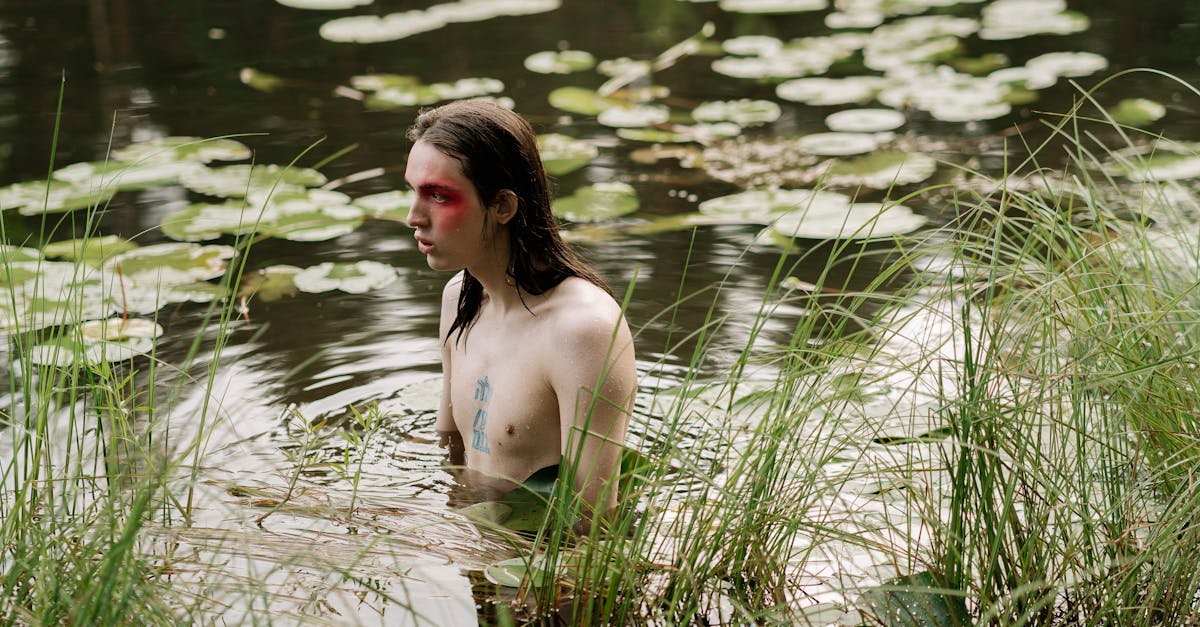 A shirtless man with painted face standing in a serene lake surrounded by nature.
