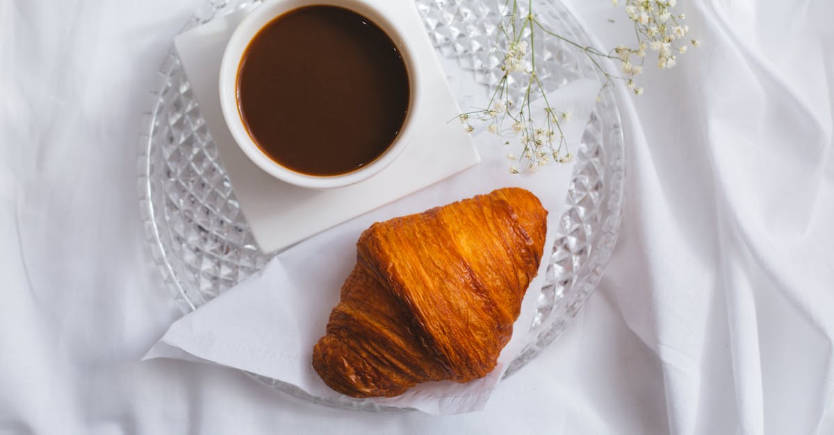 Top view of a French breakfast featuring a croissant and coffee on a glass plate, set on white linen.