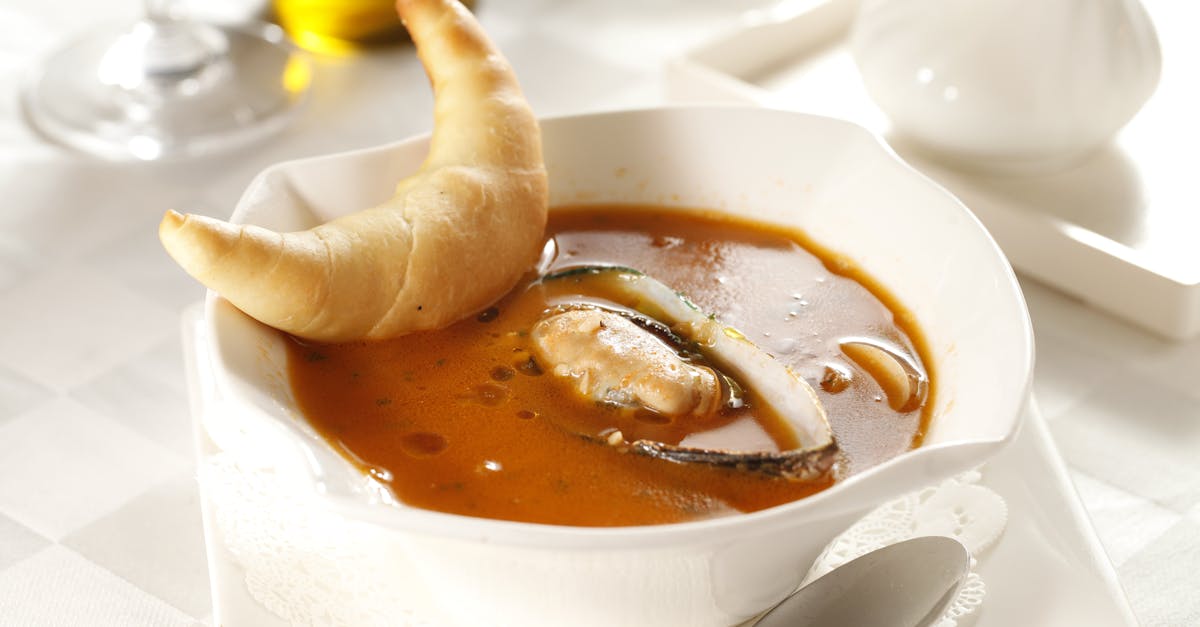 Rich tomato seafood soup with mussels served with a croissant on a white table setting.