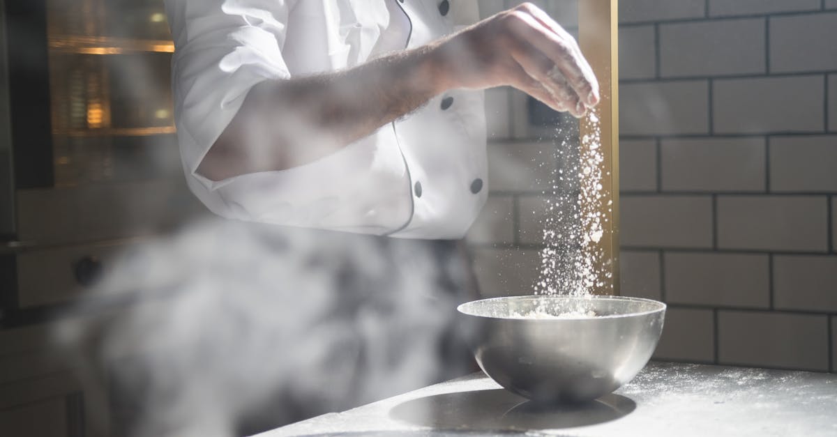 Chef in white uniform sprinkling flour into a steel bowl in a modern kitchen.